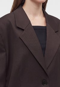Femme portant un blazer marron foncé à larges revers par-dessus un top noir côtelé, raccourci au niveau du cou et des épaules.