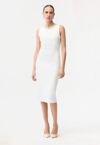 Femme debout portant une robe midi blanche sans manches avec un col orné, et des escarpins crème à bout pointu, sur un fond blanc uni.