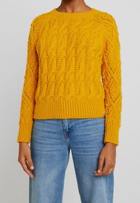 Gul stickad kabeltröja med rund halsringning, ribbade ärmslut och nederkant. Har både strukturerat mönster och matchas med ljusblåjeans.