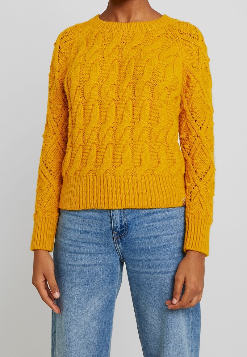 Gul stickad kabeltröja med rund halsringning, ribbade ärmslut och nederkant. Har både strukturerat mönster och matchas med ljusblåjeans.