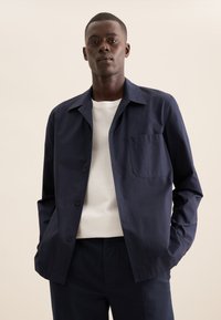 Giacca blu navy con colletto a punta, taschino sul petto singolo e una texture liscia, abbinata a una maglietta bianca sotto.