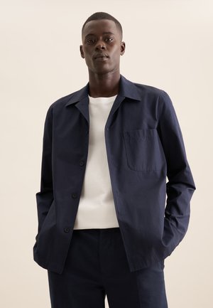 Giacca blu navy con colletto a punta, taschino sul petto singolo e una texture liscia, abbinata a una maglietta bianca sotto.