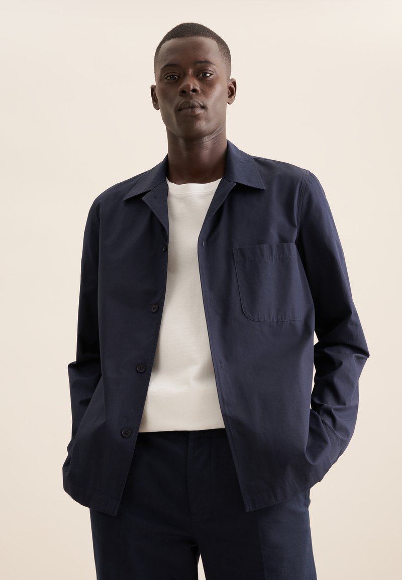 Giacca blu navy con colletto a punta, taschino sul petto singolo e una texture liscia, abbinata a una maglietta bianca sotto.