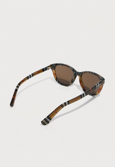 Lunettes de soleil avec verres bruns et montures à motifs ainsi que des branches dans des tons ambre, noir et blanc, inclinées sur un fond clair.