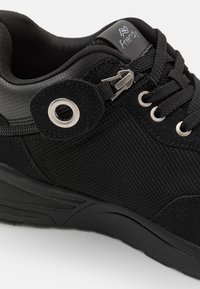 Chaussure de sport noire avec un dessus en mesh texturé, un embout renforcé, des œillets en métal et une sangle ajustable pour un maintien sécurisé.