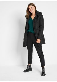 Cappotto imbottito nero con cappuccio, abbinato a un maglione verde scuro a scollo a V e pantaloni neri a sigaretta. Il modello indossa stivaletti neri con lacci.