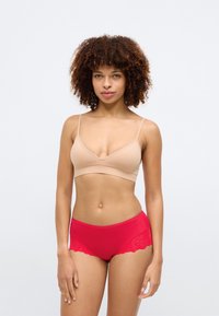 Μπεζ αόρατο σουτιέν με λεπτές τιράντες και σχέδιο σε V, συνδυασμένο με κόκκινες σλιπ που αναδεικνύουν δαντέλα με φούντες και λουλουδάτα μοτίβα.