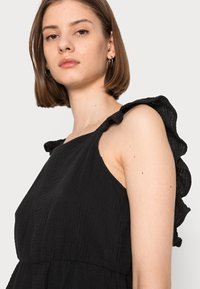 Vestido preto com alça de ombro frisada, tecido texturizado e silhueta solta. Apresenta uma cintura franzida para um detalhe adicional.