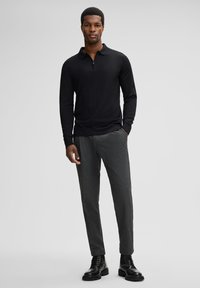 Un polo noir à manches longues avec un col zippé, associé à un pantalon gris à motifs et des bottes noires à lacets, crée un ensemble cohérent.