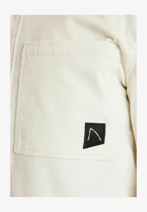 Elfenbeinfarbener Cordstoff mit einer großen Tasche und einem kleinen schwarzen Patch, der ein weißes, abstractes Liniendesign zeigt.