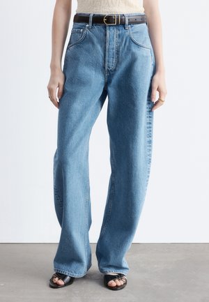 Femme portant un jean bleu clair à jambes larges avec une ceinture noire, un haut blanc et des sandales à talons ouvertes noires, debout sur un sol gris.