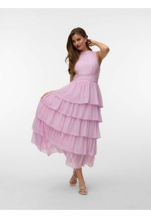 Nő ujjatlan, réteges világos rózsaszín ruhában, egyik kezével fogja a szoknyáját, a másikkal átérinti a haját, fehér háttér előtt áll.