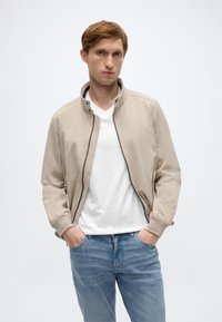 Ανοιχτό μπεζ μπουφάν τύπου bomber από λείο ύφασμα, με ψηλό γιακά, κλείσιμο με φερμουάρ και ριμπ τελείωμα στα μανίκια, σε συνδυασμό με λευκό μπλουζάκι και τζιν παντελόνι.