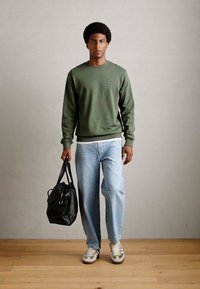 Grøn sweatshirt med rund hals, parret med lyseblå jeans og grågrønne sneakers; holder en sort lædertaske.