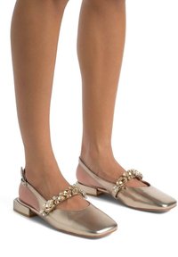 Ballerine in metallo dorato con punta quadrata, cinturino decorato con gemme trasparenti e tacco a blocco basso. Accento rosa soft sul tacco.
