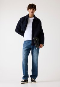 Marinefarbener Wollmischung-Hemdjacke über einem weißen T-Shirt, kombiniert mit hellblauen geraden Jeans und schwarzen Sneakers, hält eine schwarze Tasche.