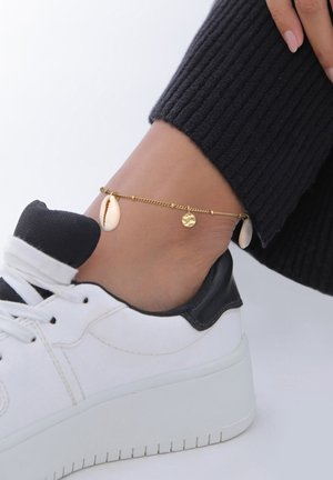 Goldenes Knöchelarmband mit Muschel- und runden Charme-Akzenten, getragen über einem weißen und schwarzen Sneaker, kombiniert mit gerippten schwarzen Socken.