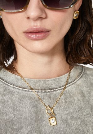 La joyería de tono dorado presenta un colgante en forma de corazón en una cadena enlazada. El modelo lleva gafas de sol y un sudadera gris con un escote redondo.