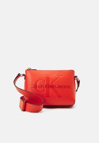 Borsa a tracolla rossa sintetica con logo "CK" in rilievo e scritta "Calvin Klein Jeans". Presenta una tracolla staccabile e una superficie liscia.