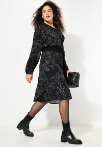 Abito nero con stampa leopardata, maniche lunghe, vita cintrata, gonna fluida. Indossato con stivali neri alla caviglia e portando una borsa nera.