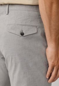 Primo piano della mano di una persona che riposa accanto a dei pantaloni a quadri grigi con un flap della tasca posteriore fissato da un bottone nero.