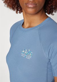 Světle modrá košile s krátkými rukávy a kulatým výstřihem, vyrobená z texturované látky a s malým vícebarevným logem na levé straně hrudi.