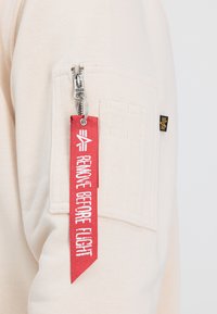 Helles beigefarbenes Sweatshirt mit einer Reißverschlusstasche, versehen mit einem roten Stoffetikett, auf dem in weißer Schrift "REMOVE BEFORE FLIGHT" steht.