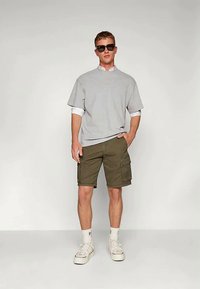 T-shirt gris surdimensionné porté par-dessus une chemise blanche à col, shorts cargo olive, baskets blanches et lunettes de soleil foncées. Tenue décontractée.