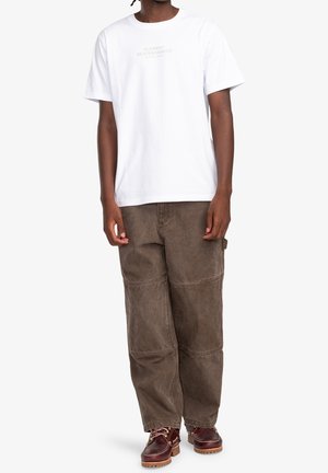 Man draagt een wit t-shirt met korte mouwen met kleine tekst, losse bruine broek met kniezomen en bruine leren laarzen met veters.