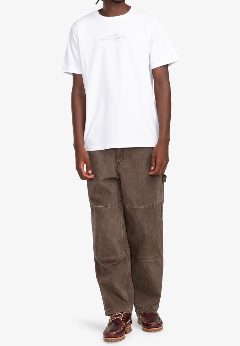 Homme portant un t-shirt blanc à manches courtes avec un petit texte, un pantalon marron ample avec des coutures aux genoux et des bottes en cuir marron avec des lacets.