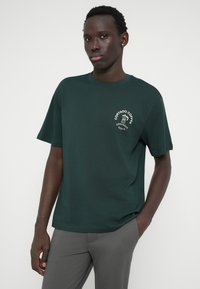 Tumed roheline puuvillane T-särk, millel on ümar kaelus ja lühikesed varrukad, ning vasakul rinnal valge graafiline trükk.