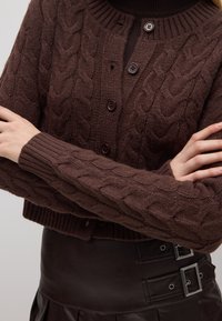 Bruine kabelgebreide cardigan met een geribde halslijn, knoopsluiting en lange mouwen. De stof oogt zacht en gestructureerd.