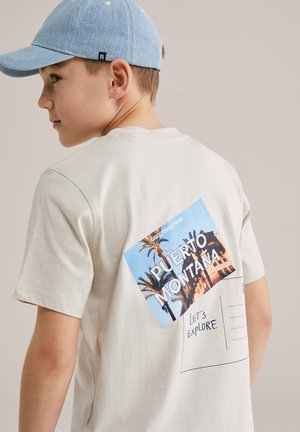 Chlapec so svetlomodrou čiapkou a krémovým tričkom s potlačou pohľadnice s nápisom "Puerto Montana" a "Let's Explore" na chrbte.