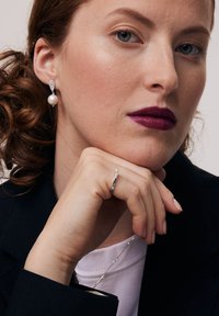 Kolczyki z akcentami perłowymi i kryształowymi, srebrny pierścionek o minimalistycznym wzornictwie, modelka w czarnym żakiecie i białej koszuli, ciemna pomadka, neutralne tło.