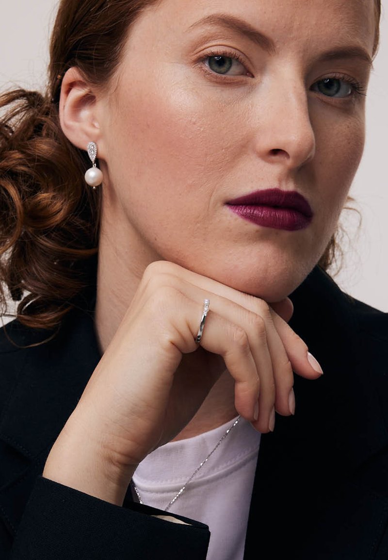 Kolczyki z akcentami perłowymi i kryształowymi, srebrny pierścionek o minimalistycznym wzornictwie, modelka w czarnym żakiecie i białej koszuli, ciemna pomadka, neutralne tło.