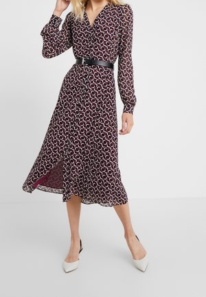 Femme portant une robe midi à manches longues à motifs, une ceinture noire et des talons hauts blancs à bout pointu, debout devant un fond uni.