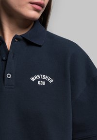 Granatowa koszulka polo z fakturowanej tkaniny, z białym haftowanym logo "WRSTBHV" i "030" na lewej części klatki piersiowej.