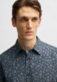 Camicia button-up di colore blu scuro con un motivo testurizzato che presenta accenti floreali di un blu chiaro. Colletto classico e chiusura a due bottoni.