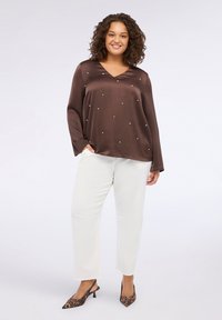 Blusa in satin marrone con scollatura a V e maniche lunghe, decorata con punti argento. Abbinata a pantaloni bianchi a vita alta e tacchi con stampa leopardata.