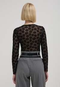 Top noir transparent à manches longues avec un motif de rose en relief, associé à un pantalon gris à rayures et une large ceinture noire.