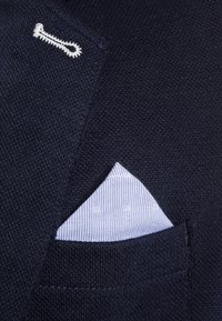Blazer in maglina blu navy con un accento bianco. Mostra un fazzoletto da taschino a righe blu e bianche che spunta dalla tasca sul petto.