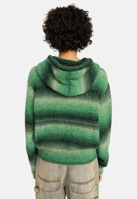 Zielony gradientowy hoodie wykonany z miękkiego, teksturowanego materiału. Posiada zaokrąglony kaptur i swobodny krój. Noszony z jasnymi dżinsowymi spodniami.