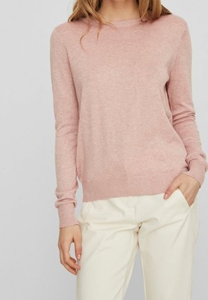 Femme portant un pull en maille rose pâle à manches longues et un pantalon blanc cassé taille haute, avec une main dans la poche et un fond neutre.