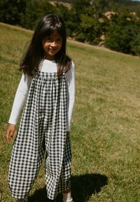 Jeune fille aux longs cheveux bruns portant une combinaison à carreaux noir et blanc et une chemise blanche à manches longues, debout dans un champ herbeux avec des arbres en arrière-plan.