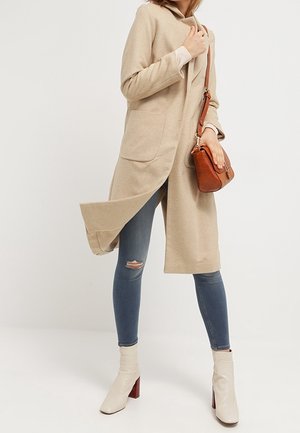Manteau beige en mélange de laine à ouverture avant, deux grandes poches, associé à un jean skinny bleu et des bottines crème à talons rouges. Sac bandoulière marron.
