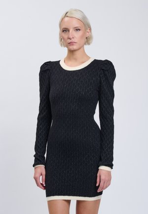 Vrouw met kort blond haar die een getailleerde zwarte jurk met lange mouwen draagt, met witte biezen aan kraag, manchetten en zoom, tegen een effen achtergrond.