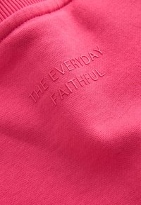 Helles pinkes Stoffmaterial mit erhabenen, auf der Oberfläche eingeprägten Texten, die "THE EVERYDAY FAITHFUL" lauten.