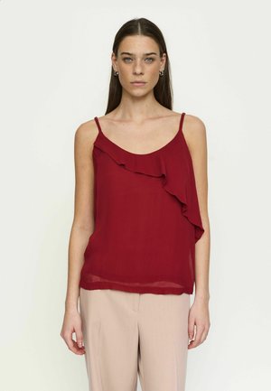 Pikkade pruunide juustega naine, kes kannab hõbedaid rõngakujulisi kõrvarõngaid, punast varrukateta toppi volangidega ja helebeeži pükse, seisab ühevärvilise tausta ees.