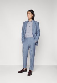 Jasnoniebieski garnitur z wyłogami, w połączeniu z wąsko paskowanym koszulem i ciemnobrązowymi butami. Spodnie są dopasowane w stylu slim fit.