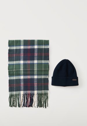 Écharpe à carreaux verts, bleu marine, blanc et rouge avec franges à côté d'un bonnet en tricot bleu marine foncé uni sur un fond clair.